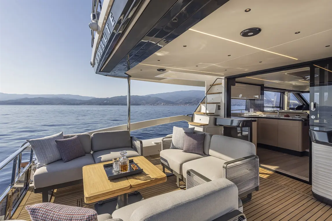 Luxury yacht deck with comfortable seating, a wooden table, and scenic sea views, featuring an adjacent indoor kitchen area.